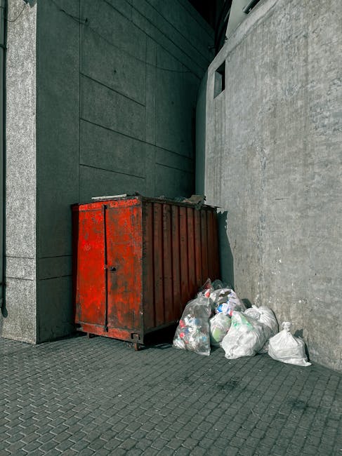 A narrow urban alleyway cluttered with various types of rubbish and debris, including large black and white garbage bags, scattered on a dark, uneven asphalt surface. In the background, there are pile