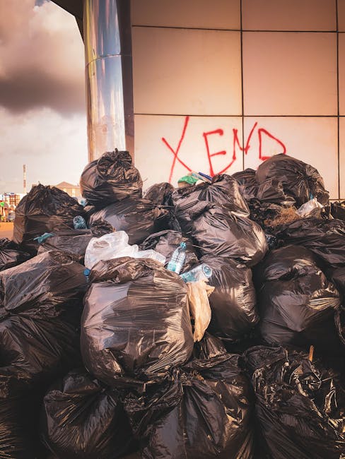 A large pile of black rubbish bags filled with waste is stacked against a modern building with tiled exterior panels. Some bags are partially torn, revealing plastic bottles and other refuse inside. T