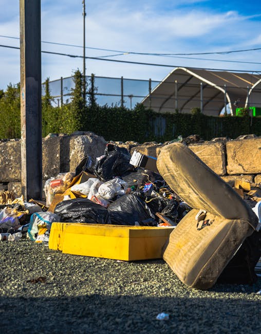 A large pile of black rubbish bags filled with waste is stacked against a modern building with tiled exterior panels. Some bags are partially torn, revealing plastic bottles and other refuse inside. T