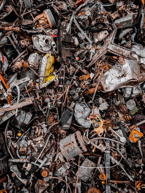 A large pile of assorted scrap metal and automotive parts, including gears, engine components, and brackets, all exhibiting rust, corrosion, and varying degrees of grime. The metals are predominantly 