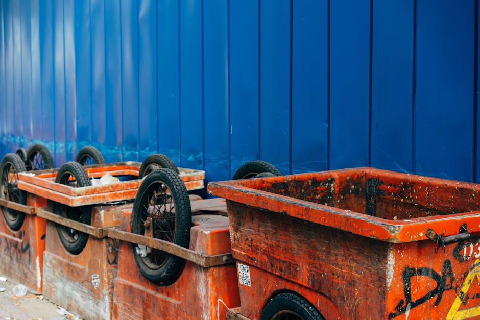 A row of large, orange plastic waste containers with black rubber wheels lined up against a blue corrugated metal fence in an urban outdoor setting. The containers show signs of heavy use, with visibl